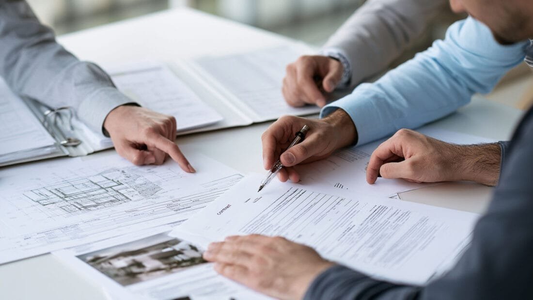 Drei Personen sitzen an einem Tisch und sehen sich Dokumente und Baupläne an und besprechen sie. Eine Person zeigt auf ein Blatt Papier, während eine andere einen Stift in der Hand hält und sich anschickt, einen Vertrag zu unterzeichnen. Die Dokumente liegen auf dem Tisch ausgebreitet.