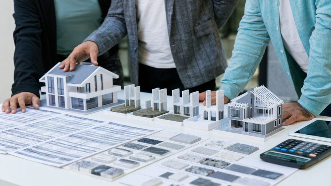 Drei Personen stehen um einen Tisch herum, auf dem sich Architekturmodelle, Baupläne, Materialmuster und ein Taschenrechner befinden. Zu sehen sind nur ihre Oberkörper und Hände.