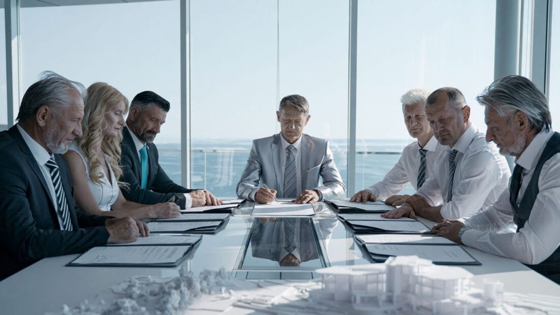 Sieben Personen in Geschäftskleidung sitzen mit Unterlagen vor sich an einem Konferenztisch und scheinen in einem modernen Büro mit großen Fenstern und Blick auf die Stadt Unterlagen zu besprechen oder durchzugehen.